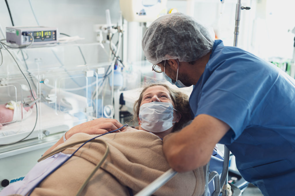 Femme avec un masque regardant son mari portant une charlotte avec le bebe en couveuse derriere