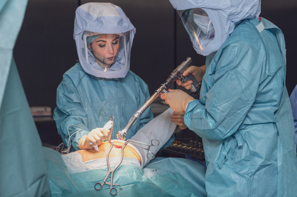 Un chirurgien habille en bleu avec son assistante tenant un outil en train d'operer la hanche d'un patient